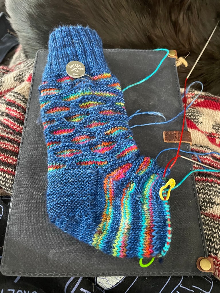 A sock on a black background. it is blue with multicolored squares that are stacked like bricks in the legs. on the foot the bricks are on the top of the foot and there are stripes on the bottom of the foot. 